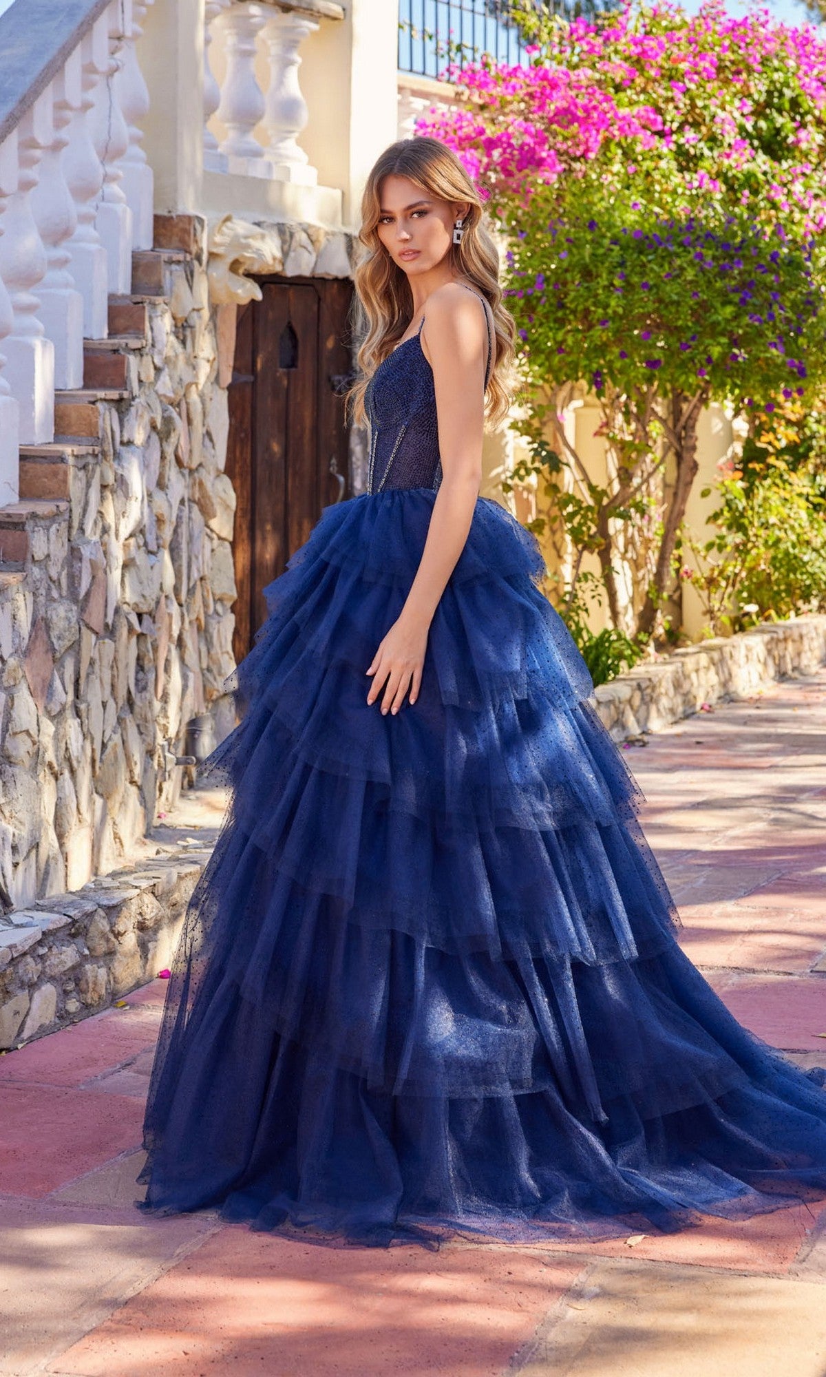 A woman in the Long Prom Dress: Juliet JT2611N, featuring a navy blue beaded corset bodice, stands outdoors on a stone path near steps and blooming bushes.